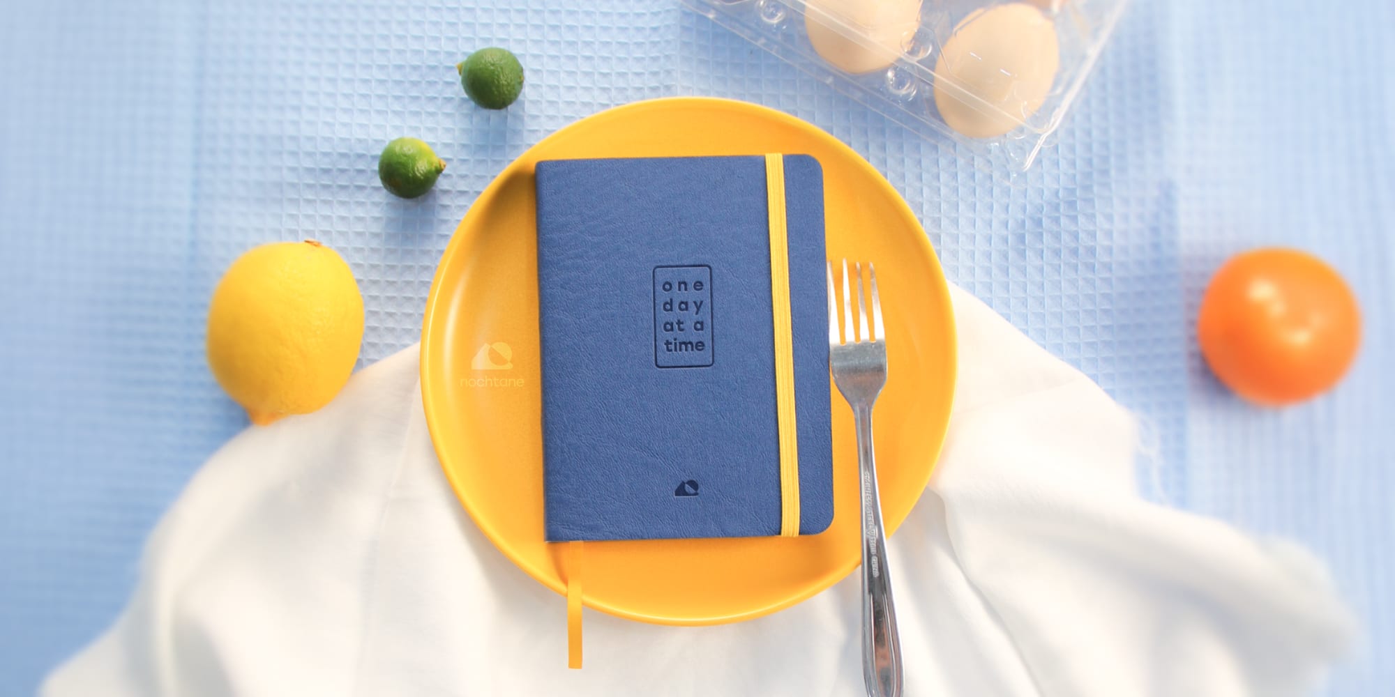 The O1 Journal, closed, on a yellow plate with a fork, surrounded by a lemon, two limes, an orange, and a carton of eggs on blue cloth.