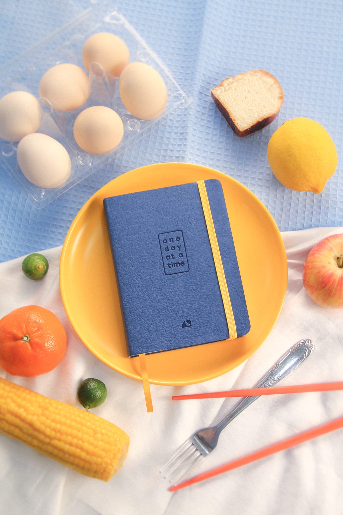 The journal on a yellow plate with a fork, among eggs, bread, corn, a lemon, an orange, and limes on blue cloth.