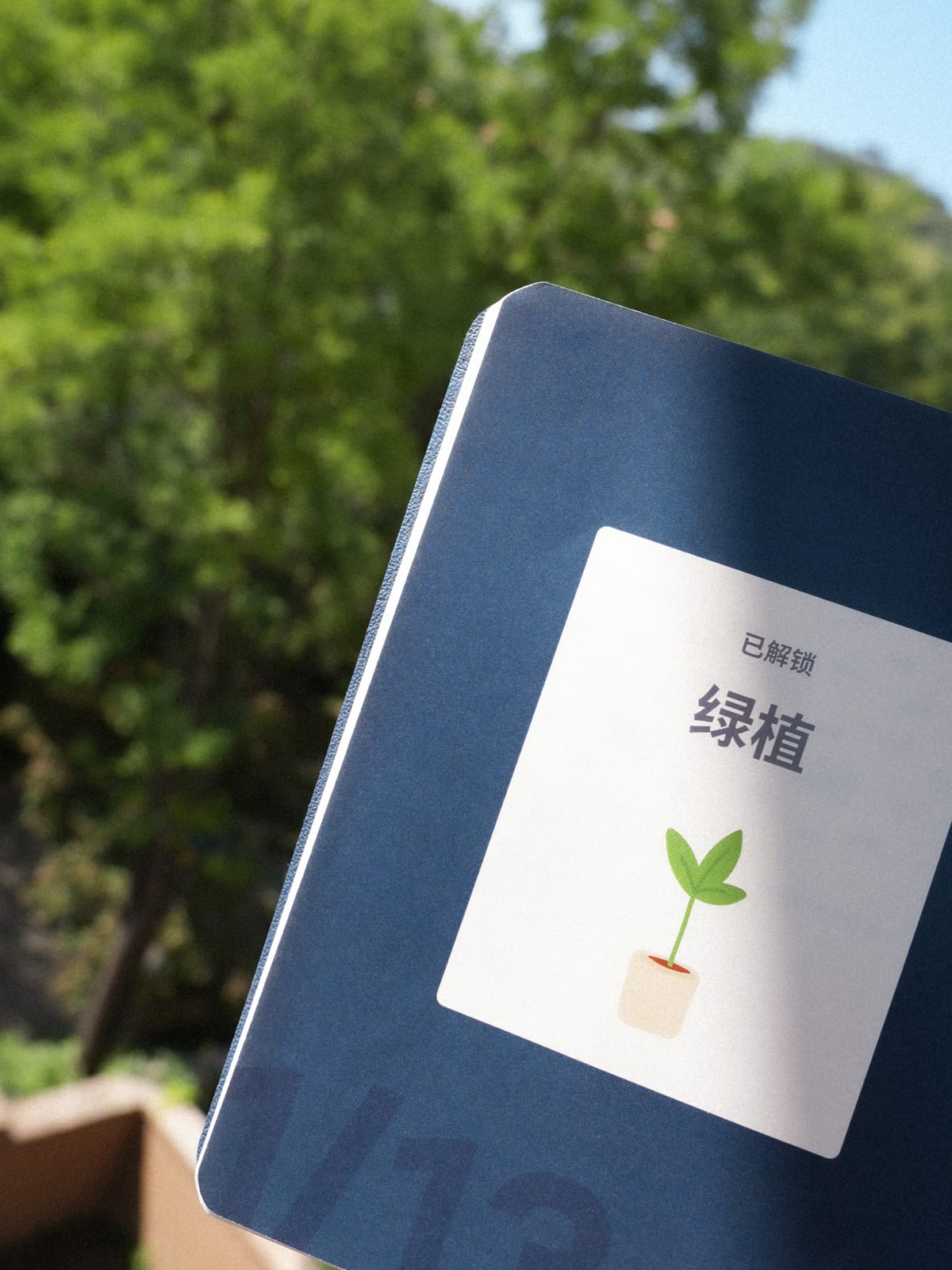 The journal held up outdoors against greenery, a green plant sticker unlocked on its cover.