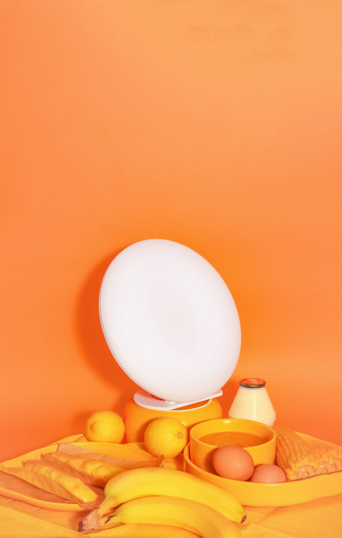 The lamp lit on an orange backdrop, among bananas, lemons, eggs, and bread.