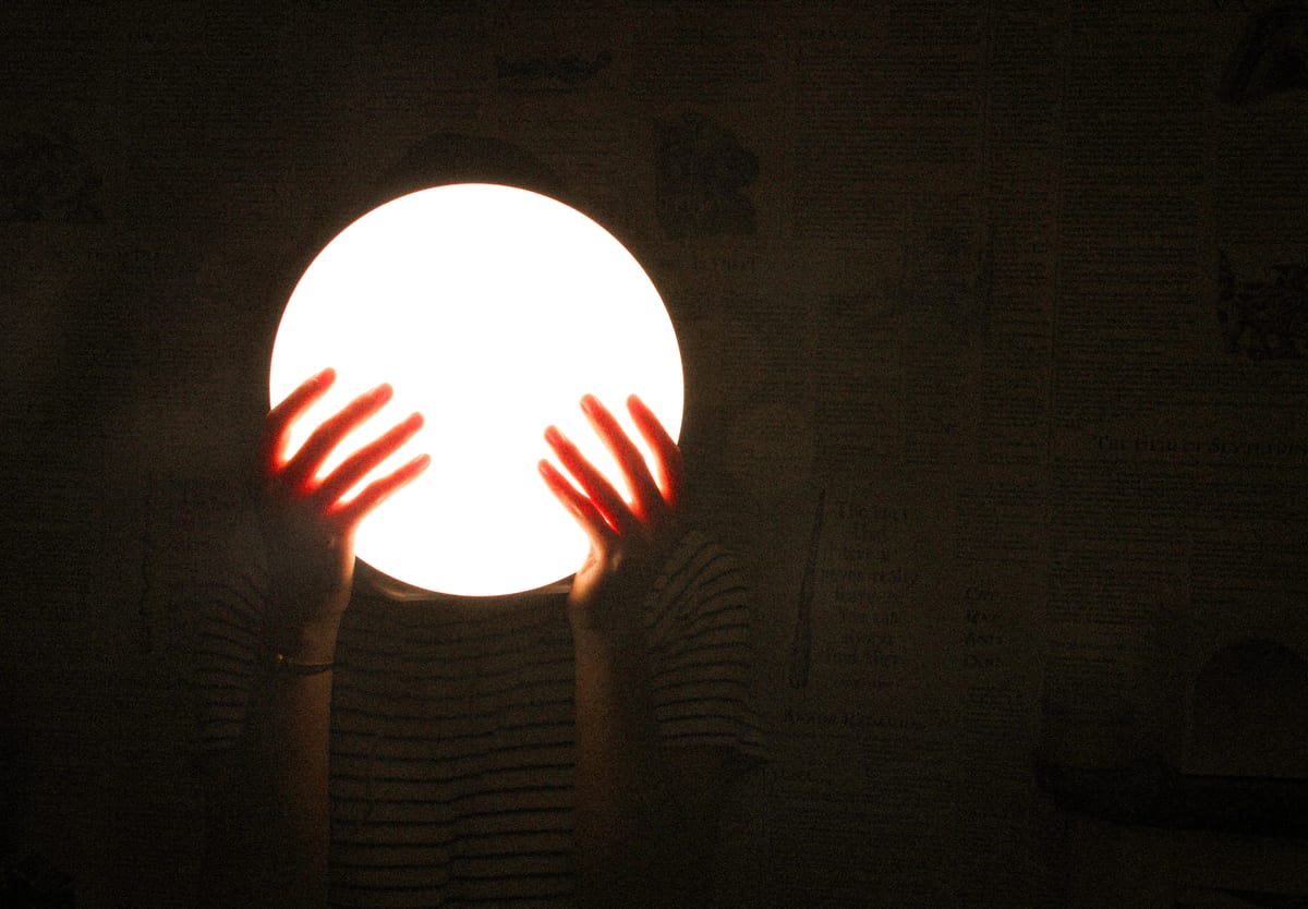 Two hands around the lit lamp in the dark, the fingers glowing red where the light passes through them.