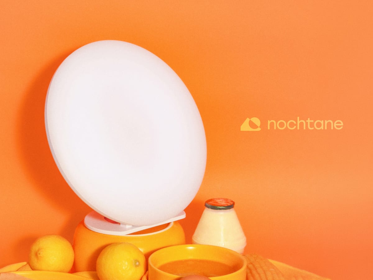 The O1 Sun Lamp lit on an orange backdrop, among lemons, a bowl of juice, and a bottle of yogurt drink, the nochtane lockup at the right.