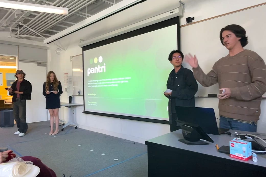 Four team members presenting Pantri in a classroom. Two stand at a desk mid-sentence, two wait beside the screen holding notes, and the title slide behind them shows the pantri wordmark in white on green above three dots.